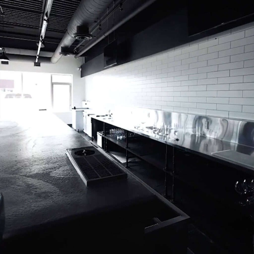 Iron Shake Inc A modern, empty commercial kitchen by Iron Shake Inc., featuring stainless steel counters, white subway tile walls, and industrial ceiling fixtures—perfectly crafted with expert commercial construction services and brightened by daylight from a large front window. Commercial Contractor