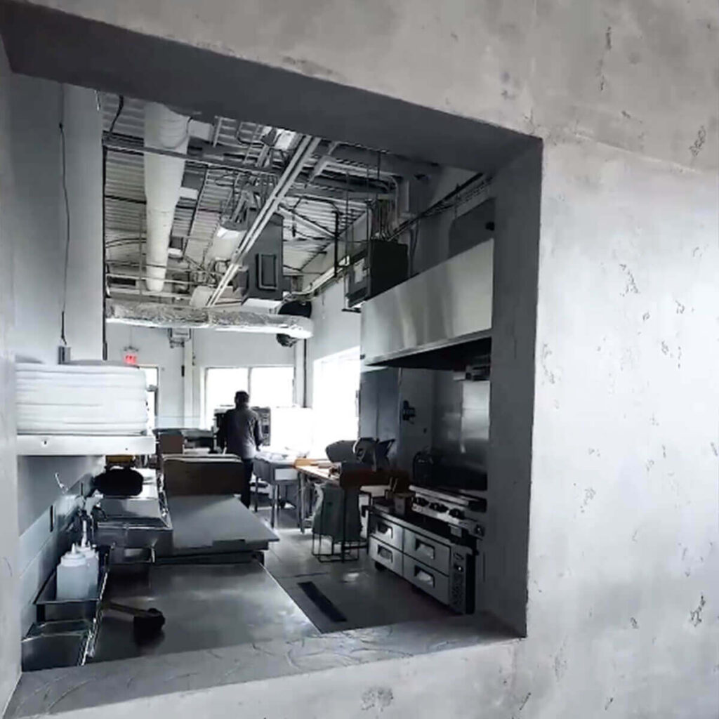 Iron Shake Inc View through a window into an industrial kitchen with stainless steel equipment, tables, and a person near the back—showcasing the quality work of Iron Shake Inc, experts in commercial construction services. Commercial Contractor