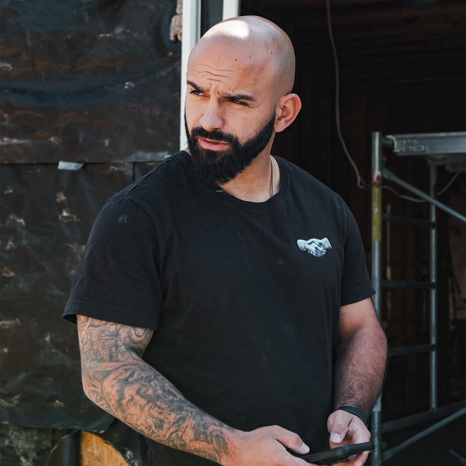 Iron Shake Inc A bald man with a beard and tattooed arms stands holding a phone, wearing a black t-shirt, in front of a partially constructed building, representing Iron Shake Inc’s expertise in commercial construction services. Commercial Contractor