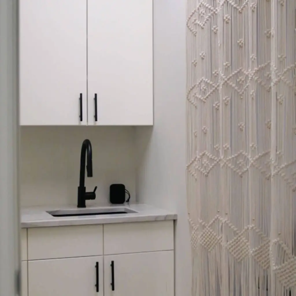 Iron Shake Inc A small kitchenette with white cabinets, a black faucet, and a countertop sits beside a decorative cream-colored macramé curtain—crafted with the quality you’d expect from Iron Shake Inc’s commercial contracting expertise. Commercial Contractor