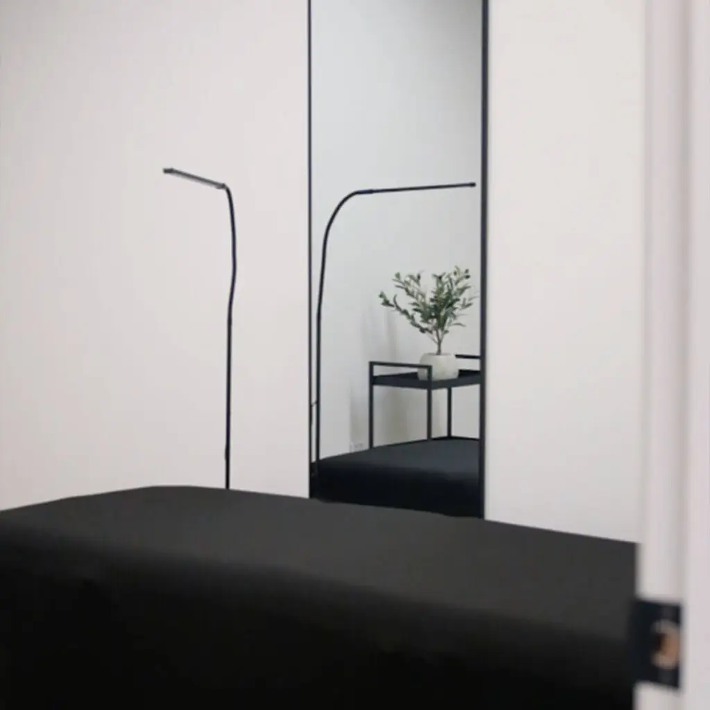 Iron Shake Inc A minimal room with a black table, a tall mirror reflecting a potted plant on a black stand, and a thin black floor lamp against white walls—crafted with the precision of Iron Shake Inc.'s commercial construction services. Commercial Contractor