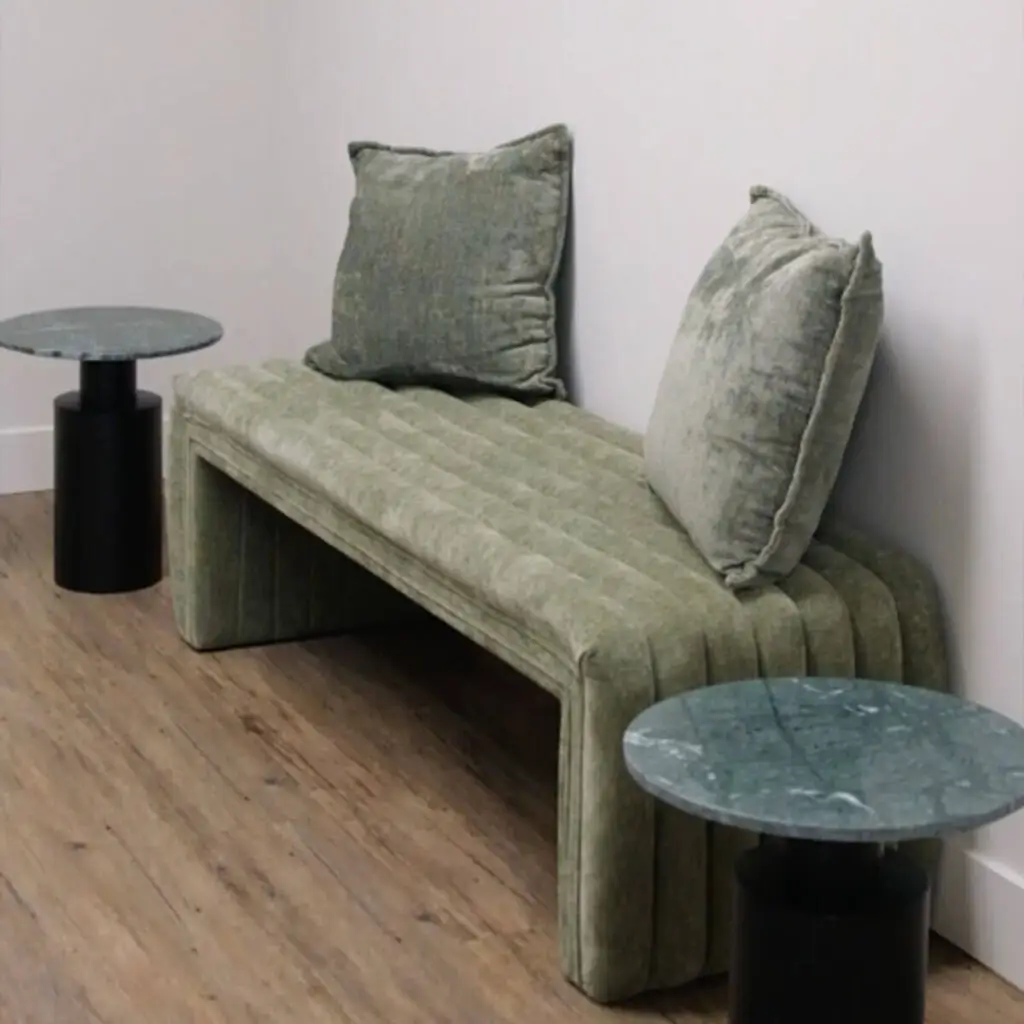 Iron Shake Inc A green upholstered bench with two matching cushions is positioned against a white wall, flanked by two round marble-top side tables on a wood floor—a stylish look achieved with the expertise of Iron Shake Inc's commercial construction services. Commercial Contractor